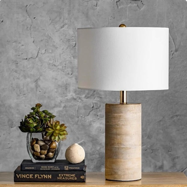 Lampe de chevet Lumeoo avec pied cylindrique en bois texturé et abat-jour blanc, posée sur des livres près de plantes.