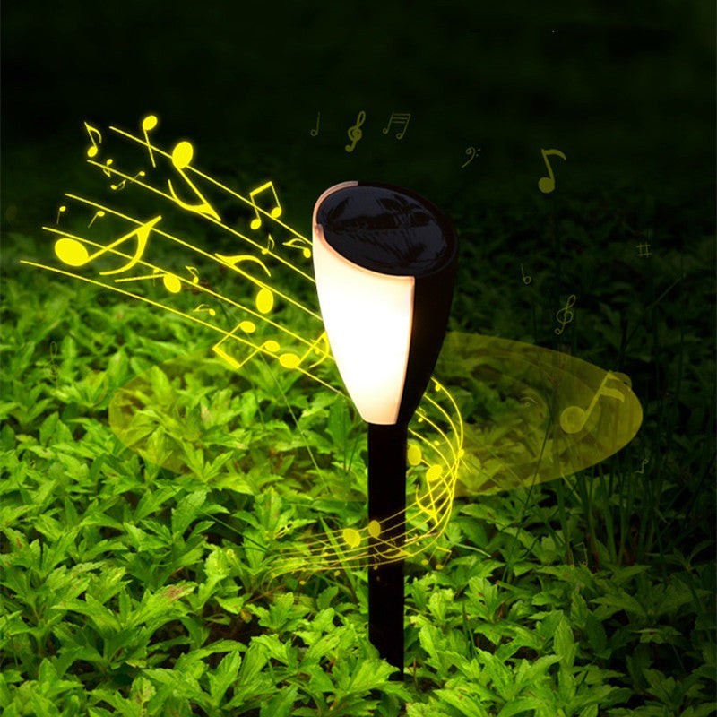 Borne solaire Lumeoo SONOVA bicolore avec haut-parleur intégré diffusant de la musique dans un jardin verdoyant.