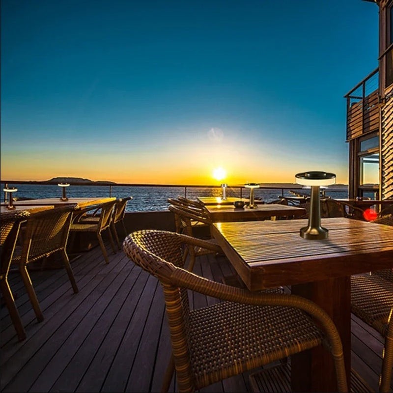 Lampe de table sans fil Lumeoo dorée posée sur une table en bois en terrasse face à un coucher de soleil sur la mer