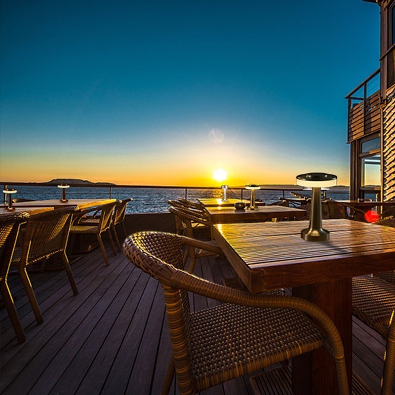 Lampe de table sans fil Lumeoo en métal noir sur une terrasse en bois au coucher du soleil, design LED dimmable.